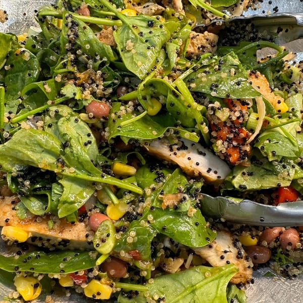 Close-up of a healthy salad with spinach, quinoa, chicken, corn, and beans, with serving tongs.